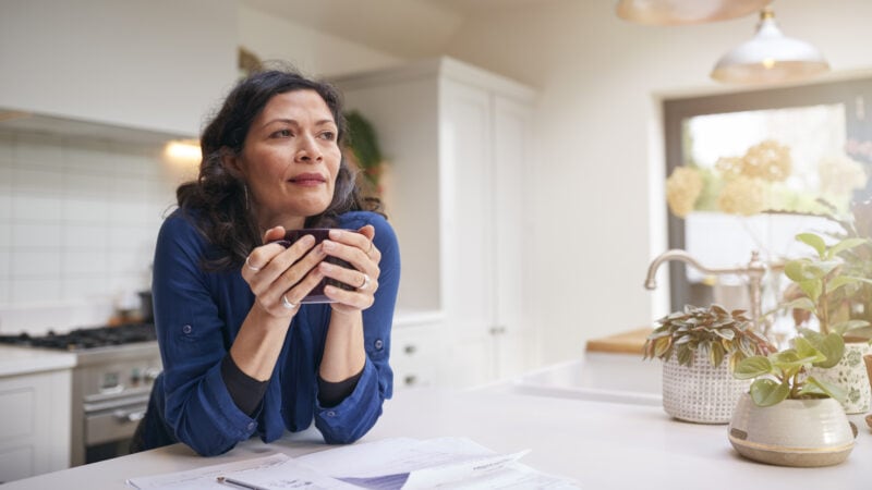 Thoughtful Mature Woman Reviewing Domestic Finances And Paperwork Drinking Coffee In Kitchen At Home