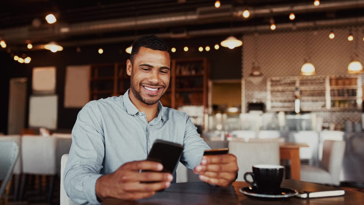 Happy man holding mobile phone typing card data to make online payment sitting in cafe