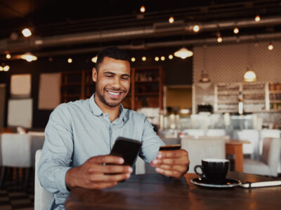 Happy man holding mobile phone typing card data to make online payment sitting in cafe