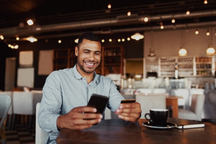 Happy man holding mobile phone typing card data to make online payment sitting in cafe